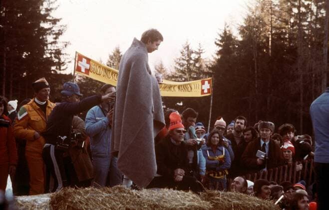 Manfred Schumann (BR Deutschland I) in eine wärmende Decke gehüllt und auf einem Strohhaufen vor den Fans Bobsport OS Winter Herren Olympische Winterspiele Innsbruck 1976, Olympia, Olympiade, Spiele, Bob, Zweierbob, Zweier, Vdia Totale Igls