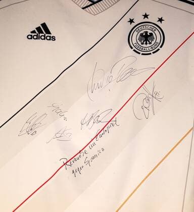 FUSSBALL DEUTSCHE NATIONALMANNSCHAFT 2011 Nach der Präsentation signierten u.A. Lukas Podolski, Holger Badstuber, Andre Schürrle und Mats Hummels ein Trikot. Einer der Spieler hat REVANCHE IM ENDSPIEL GEGEN SPANIEN draufgeschrieben.  o0 Objekte Equipment Trikot Nationaltrikot Detail Logo Verbandslogo Unterschrift Signatur Autogramm