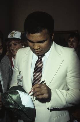 Muhammad Ali (Ex-Boxprofi) 09 84 wot Mann ehemaliger Boxer Box-Profi Profi-Sport Profisport Boxen Weltmeister Boxweltmeister dunkelhäutig dunkelhaarig kurzhaarig seriös Jackett Hemd weiss Krawatte stehend Saal Halle halb hoch nach unten schauend Stift haltend schreibend signierend autogrammegebend Autogramme gebend auf Kappe Baseballkappe unterschreibend Menschen Fans Cassius Clay
