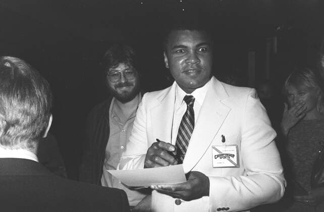 Muhammad Ali (re., Ex-Boxprofi) 09 84 wot Mann ehemaliger Boxer Box-Profi Profi-Sport Profisport Boxen Weltmeister Boxweltmeister dunkelhäutig dunkelhaarig kurzhaarig seriös Jackett Hemd weiss Krawatte stehend Saal Halle halb quer fragend lächelnd aufschauend zur Seite schauend Stift haltend Unterlagen Zettel Menschen Fans Cassius Clay