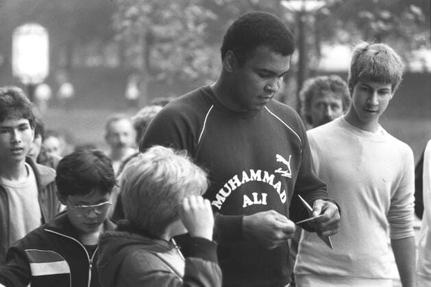 Muhammad Ali (Ex-Boxprofi) 09 84 wot Mann ehemaliger Boxer Box-Profi Profi-Sport Profisport Boxen Weltmeister Boxweltmeister dunkelhäutig dunkelhaarig kurzhaarig leger Sportzeug Sportanzug Jogginganzug blau dunkel dunkelblau Aufschrift Aufdruck Muhammad Ali Name Marke Puma Turnschuhe stehend draussen halb quer lächelnd nach unten schauend Unterlagen Zettel haltend Brief öffnend um ihn herum Menschen Fans Kinder Jugendliche Cassius Clay