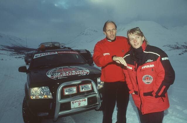 Jutta Kleinschmidt und Hans-Joachim Stuck im Yukon- Territorium (Kanada) 02 00 mapo Motorsportlerin Motorsport Auto Autosport Rallyes Rallye Rallyesport Rallyefahrerin Rallyefahrer Fahrerin Fahrer leger sportlich rot-schwarz stehend draussen vor Auto PKW Wagen Logo Fulda halb quer lächelnd Gespräch sich umschauend etwas beobachtend suchend