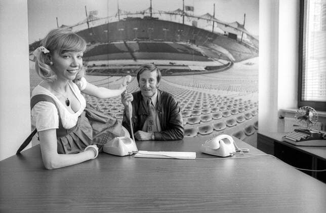 Rudi Gutendorf (BR Deutschland) sitzt vor einem Bild des Münchner Olympiastadions an einem Tisch, auf dem sich eine Frau im Dirndl rekelt