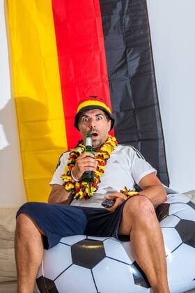 Football fan with fan merchandise drinks beer from bottle and looks at Germany game on TV