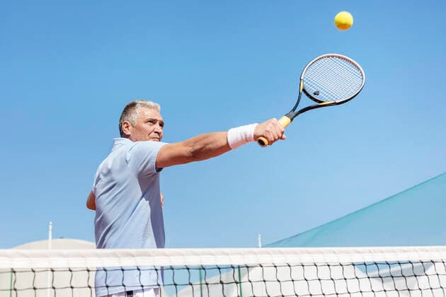Low angle view of confident mature man hitting tennis ball with racket on court against clear blue sky INH_19064_13251