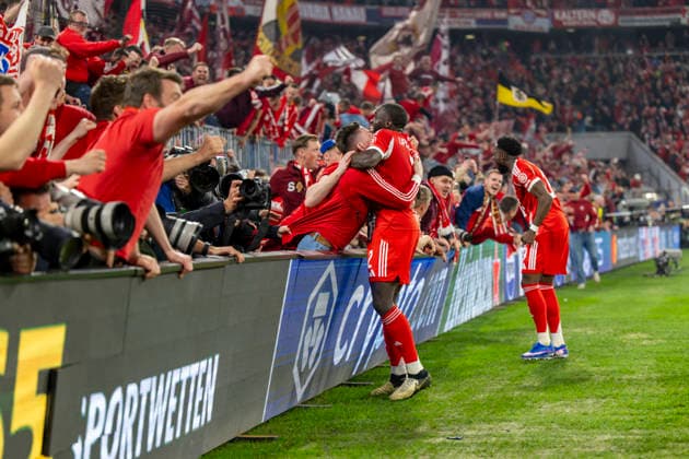 15 04 2026, Football UEFA Champions League 2025 2026 quarter-final second leg, FC Bayern Munich Real Madrid, in the Allianz Arena Munich, fans storm the interior before the final whistle and push over the boards with the photographers while celebrating the goal, two photographers are injured, even unconscious, and have to be treated by a doctor afterwards Dayot Upamecano FC Bayern Munich hugs a fan