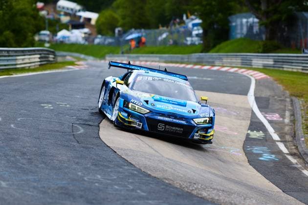 The Audi R8 LMS GT3 with starting number 16 from Scherer Sport PHX negotiates a tight bend on the Nordschleife of the Nürburgring The blue and yellow car bears the sponsors HYLO, TÜV Rheinland and H R and is fitted with Michelin tires The characteristic crash barriers and the green surroundings of the Eifel can be seen in the background Alexander Sims Ben Green Christopher Haase Scherer Sport PHX Audi R8 LMS GT3 16 from Scherer Sport PHX on the Nordschleife Copyright: xspeedshotx