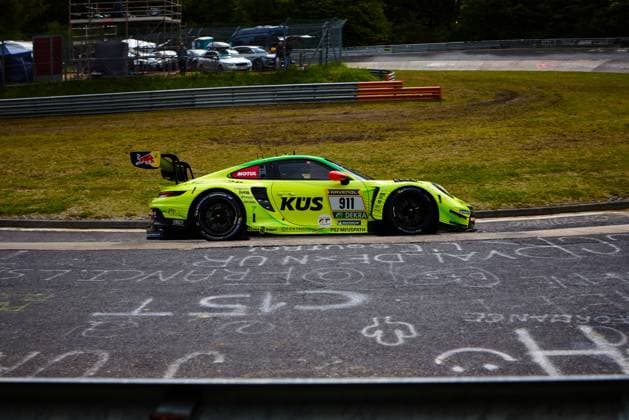 The bright yellow and green Porsche 911 GT3 R with the starting number 911 of the Manthey team drives around the Nürburgring race track The strikingly designed vehicle Ayhancan Güven Kevin Estre Thomas Preining Manthey bears the main sponsors KÜS and RAVENOL prominently on the bodywork as well as a Red Bull sticker on the rear wing, crash barriers and vehicles in the paddock can be seen in the background, while fan lettering can be read on the asphalt in the foreground Porsche 911 GT3 R 911 from Manthey racing at the Nürburgring Copyright: xspeedshotx