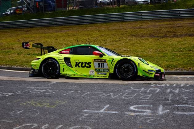 The Porsche 911 GT3 R with the starting number 911 of the Manthey team drives through a bend at the Nürburgring The car Ayhancan Güven Kevin Estre Thomas Preining Manthey presents itself in eye-catching neon yellow green with the KÜS main sponsorship and the logos of RAVENOL, DEKRA, Motul and Red Bull logos On the asphalt, fan lettering in chalk underlines the atmosphere of the 24-hour race Porsche 911 GT3 R 911 from Manthey in KÜS design at the Nürburgring Copyright: xspeedshotx