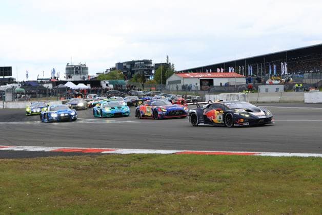 The Lamborghini Huracán GT3 with the starting number 84 of the Red Bull Team ABT leads the field at the start of the ADAC RAVENOL 24h Nürburgring Luca Engstler Mirko Bortolotti Patric Niederhauser Red Bull Team ABT is in front in the first corner, followed by numerous other GT3 cars - including a Mercedes AMG GT3, a turquoise-colored Lamborghini and several Audi R8 LMSs - are pushing towards the Nordschleife on the starting grid The packed grandstand and the paddock infrastructure of the Nürburgring can be seen in the background Race start at the 24h Nürburgring 84 Red Bull Team ABT leads the field Copyright: xspeedshotx