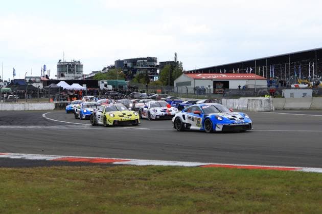 In the foreground, the blue and white Porsche 911 GT3 with the starting number 918 from Mühlner Motorsport powered by H R leads the field through a tight bend on the Nürburgring Grand Prix circuit The yellow and black Porsche 911 GT3 with the number 992 follows closely behind, followed by other GT cars in a tight group The main grandstand and the AMG logo on the Nürburgring control tower can be seen in the background The picture shows the intense opening phase of the ADAC RAVENOL 24h Nürburgring 2026 in cloudy weather Starting phase at the 24h Nürburgring - Porsche 918 leads the field Copyright: xspeedshotx