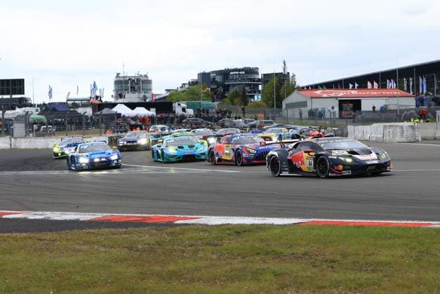 The Lamborghini Huracán GT3 with starting number 84 from Red Bull Team ABT leads a dense starting field in a corner of the Nürburgring In the background, numerous other GT3 vehicles can be seen fighting for positions, including a blue Audi, a turquoise Lamborghini and a Mercedes AMG GT3 Spectators line the track and the Nürburgring infrastructure with the AMG hospitality area is clearly visible in the background Luca Engstler Mirko Bortolotti Patric Niederhauser Red Bull Team ABT Lamborghini 84 Red Bull ABT leads the field at the Nürburgring Copyright: xspeedshotx