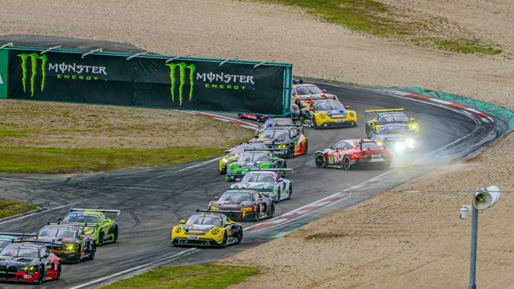 A large field of drivers battles close together through a bend at the Nürburgring Numerous GT vehicles from various brands and teams can be seen crowded together on the track, including several Porsche 911 GT3s and a striking red vehicle Monster Energy advertising banners line the track boundary The image conveys the intense atmosphere of the 24-hour race at the Nürburgring Dense field of drivers in the bend at the Nürburgring 24 Hours 2026 Copyright: xMichaelxSchäferx