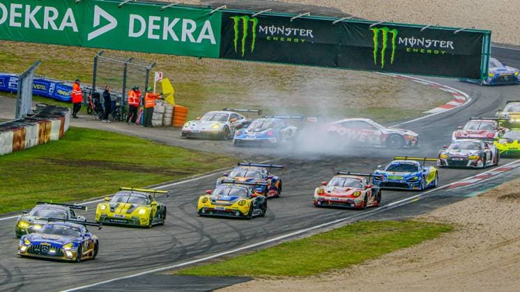 Dramatic scene in the starting phase of the ADAC RAVENOL 24h Nürburgring 2026 Numerous GT3 cars take a tight bend while a car in the midfield is struggling with locked wheels and heavy smoke Track marshals in orange safety vests display yellow flags A blue and gold Mercedes AMG GT3 can be seen in the foreground, accompanied by several yellow Porsche 911 GT3 R Monster Energy and DEKRA are prominently visible in the background as trackside advertising Starting phase with incident at the 24h Nürburgring 2026 Copyright: xMichaelxSchäferx