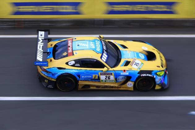 The Mercedes AMG GT3 with starting number 26 from PROsport Racing drives through the pit lane at the Nürburgring The vehicle Adam Christodoulou Christopher Lulham Marek Böckmann Mikaël Grenier PROsport Racing presents itself in an eye-catching yellow and blue pixel design with sponsors such as RAVENOL, Bilstein, HELLA, DEKRA, WEMPE and Falken The AMG rear wing is clearly visible as the vehicle glides through the pit lane with a slight blur of movement Mercedes AMG GT3 26 from PROsport Racing in the pit lane Copyright: xspeedshotx