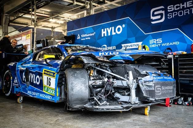 Der Alexander Sims & Ben Green & Christopher Haase (Scherer PHX) von Scherer Sport PHX steht nach einem schweren Unfall in der Garage am Nürburgring. Die Front des blauen Audi R8 GT3 ist vollständig demoliert – Karosserieteile fehlen, der Frontbereich ist offen, Kabel und Strukturteile liegen frei. Der Unterboden und die Aerodynamik-Komponenten sind stark deformiert. Im Hintergrund ist die Teamwand mit den Sponsoren HYLO, RAVENOL, TÜV Rheinland und IRS Dent Wizard sichtbar. Ein Teammitglied beobachtet das Fahrzeug im Hintergrund. Schwer beschädigter Audi R8 GT3 16 von Scherer Sport PHX in der Box *** The Alexander Sims Ben Green Christopher Haase Scherer Sport PHX from Scherer Sport PHX is in the garage at the Nürburgring after a serious accident The front of the blue Audi R8 GT3 is completely demolished - body parts are missing, the front area is open, Cables and structural parts are exposed The underbody and the aerodynamic components are heavily deformed The team wall with the sponsors HYLO, RAVENOL, TÜV Rheinland and IRS Dent Wizard is visible in the background A team member observes the vehicle in the background Heavily damaged Audi R8 GT3 16 from Scherer Sport PHX in the pits Copyright: xMichaelxGelhardtx