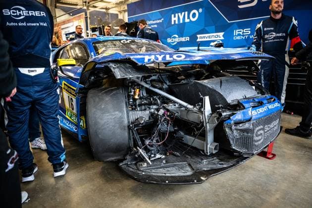 The Alexander Sims Ben Green Christopher Haase Scherer Sport PHX from Scherer Sport PHX is parked in the pit garage at the Nürburgring after a serious accident The front of the blue GT3 car is completely destroyed Body parts are missing, radiator pipes, cables and chassis components are exposed The front section is so badly deformed that the car barely appears to be fit to race, Team members in blue Scherer overalls stand around the vehicle Sponsors such as HYLO, RAVENOL, IRS Dent Wizard, DEKRA and Cam Shaft are still recognizable on the heavily damaged vehicle Heavily damaged GT3 16 from Scherer Sport PHX in the pits Copyright: xMichaelxGelhardtx