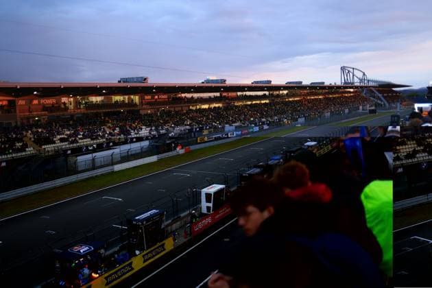 The main grandstand at the Nürburgring is packed at the ADAC RAVENOL 24h Nürburgring 2026 At dusk, the BMW M Power advertising banners in the covered grandstand light up as thousands of spectators watch the race action In the background, the characteristic roller coaster construction of the amusement park and the cloudy evening sky over the Eifel can be seen Fully occupied main grandstand at the Nürburgring at dusk Copyright: xspeedshotx