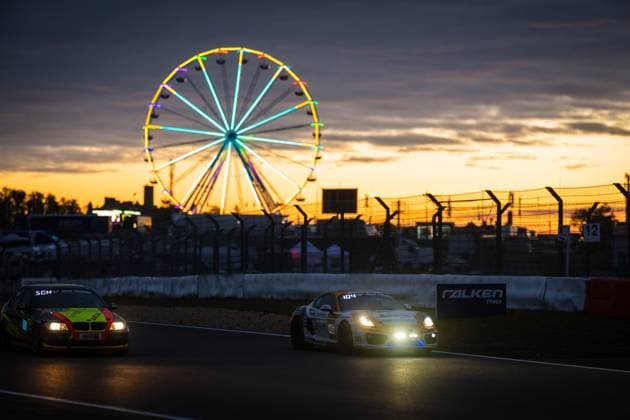Two cars cross the track at the Nürburgring at an impressive sunset A white Porsche Cayman with the number 104 in the foreground, followed by a yellow and red BMW In the background, the colorful Ferris wheel of the paddock shines against the orange and purple evening sky A Falken Tyres banner can be seen on the track boundary, the headlights of the vehicles illuminate the twilight Dramatic sunset atmosphere at the Nürburgring with Ferris wheel Copyright: xMichaelxGelhardtx