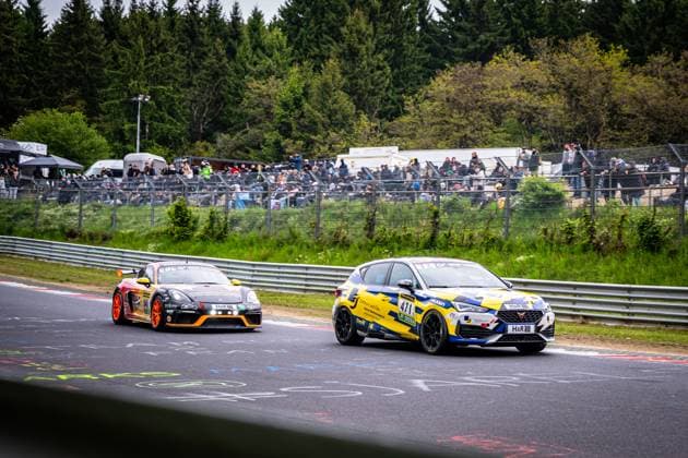 Two cars engage in a close duel on the Nürburgring at the ADAC RAVENOL 24h Nürburgring 2026 In the foreground, Andreas Winterwerber Michael Eichhorn Tony Roma Jung Motorsport - a CUPRA Leon Competición in yellow and blue design with H R and Falken sponsorship - drives along the track Behind it follows a black and orange Porsche 718 Cayman GT4 with starting number 441 and H R sponsorship Numerous spectators follow the race from the grandstand behind the crash barrier, surrounded by the typical green Eifel landscape CUPRA 471 and Porsche 441 battle it out on the Nürburgring Copyright: xMichaelxGelhardtx