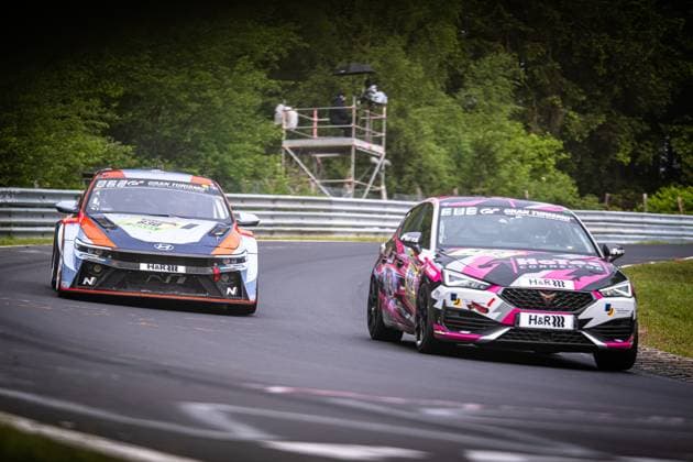 Two cars fight a close duel on the Nordschleife of the Nürburgring On the left, a Hyundai Elantra N TCR in blue and orange design with H R sponsoring and Gran Turismo branding fights for position On the right, a CUPRA Leon in an eye-catching pink, black and white camouflage design, also with H R sponsoring and Gran Turismo branding Both cars drive through a fast corner on the green forest section of the Nordschleife The start numbers of both cars are not clearly legible due to the driving and the angle of the picture Hyundai and CUPRA in a duel on the Nordschleife Copyright: xMichaelxGelhardtx