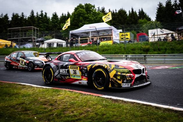 The BMW Z4 GT3 Christian Scherer Henry Walkenhorst Julian Reeh Ralf Schall Saugmotoren Motorsport 3M from Saugmotoren Motorsport 3M takes a corner on the Nordschleife at the Nürburgring The car wears a striking red, black and yellow livery with sponsors such as W S T Welding Specialists Team, RAVENOL, DEKRA and 3M Another car with starting number 100 follows in the background, while spectators and team tents line the track BMW Z4 GT3 36 from Saugmotoren Motorsport 3M on the Nordschleife Copyright: xMichaelxGelhardtx