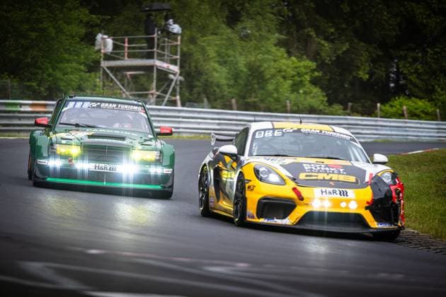 Two cars battle it out on the wet Nordschleife of the Nürburgring A yellow and black Porsche 718 Cayman GT4 with CMB and H R sponsorship is in the foreground, while a dark green racing car with PARKSIDE and H R sponsorship and Gran Turismo branding is close behind it The track is damp, with a green forest edge with a track marshals scaffolding visible in the background Porsche 718 Cayman GT4 and green racing car on the Nordschleife Copyright: xMichaelxGelhardtx