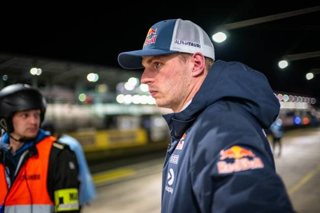 Max Verstappen stands concentrated in the illuminated pit lane of the Nürburgring He wears a Red Bull cap with AlphaTauri lettering and a dark blue Red Bull jacket with Viaplay and SOTO sponsors A marshal in an orange safety vest can be seen in the background The nocturnal atmosphere of the pit lane is characterized by the bright hall lighting Max Verstappen in the pit lane at the 24h Nürburgring 2026 Copyright: xMichaelxGelhardtx