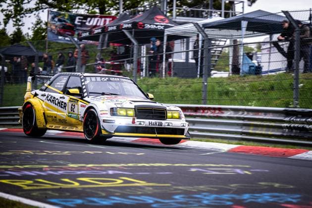 The spectacular Mercedes Benz 190E with starting number 62 from HWA Engineering Speed takes a bend on the Nürburgrings Nordschleife with impressive dynamism The yellow and white vehicle bears the sponsors Kärcher, RAVENOL, SONAX, H R and Hankook and features Gran Turismo branding Spectators behind the safety fence and a 24MX tent can be seen in the background The switched-on headlights and the characteristic rear spoiler emphasize the racing character of the historic vehicle Luca Ludwig Markus Winkelhock Sebastian Asch HWA Engineering Speed Mercedes Benz 190E 62 from HWA Engineering Speed on the Nordschleife Copyright: xMichaelxGelhardtx