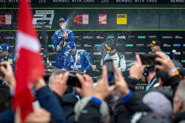 Christian Krognes stands on the podium and is sprayed with champagne while photographers and fans document the award ceremony Sponsor banners with RAVENOL, ADAC and other partners are visible in the background Award ceremony on the podium of the 24h Nürburgring 2026 Copyright: xMichaelxGelhardtx