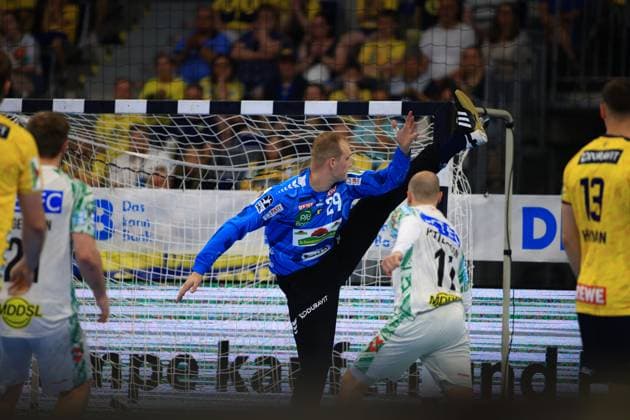 David Späth 29 Rhein Neckar Löwen, wearing a blue goalkeepers jersey, stretches into the goal of the SAP Arena with a high leg lift to make a save In the foreground are players from both teams, including an SC Magdeburg player in a white jersey and Löwen player 13 Sebastian Heymann in a yellow jersey David Späth 29 Rhein Neckar Löwen making a spectacular save Copyright: xspeedshotx