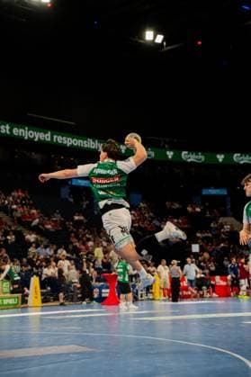 A handball player in a green jersey with sponsors performs a jump shot and is completely lifted off the ground The Barclays Arena court with spectators in the background is clearly visible Players in green jerseys performing a jump shot at Barclays Arena Copyright: xPhilippHeistx