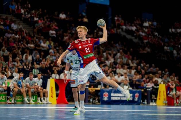 Frederik Bo Andersen wears the red jersey of HSV Hamburg and performs a jump shot while holding the ball above head height In the background you can see players of Frisch Auf Göppingen in white jerseys and the spectators of the Barclays Arena Handball Sport Verein Hamburg vs Frisch Auf Göppingen Copyright: xPhilippHeistx