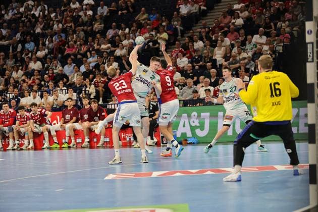 Oskar Neudeck in white Göppingen jersey throws the ball from a raised position against the red Hamburg defenders The Barclays Arena is packed with spectators Oskar Neudeck 2 Frisch Auf Göppingen throwing against HSV Hamburg Copyright: xspeedshotx
