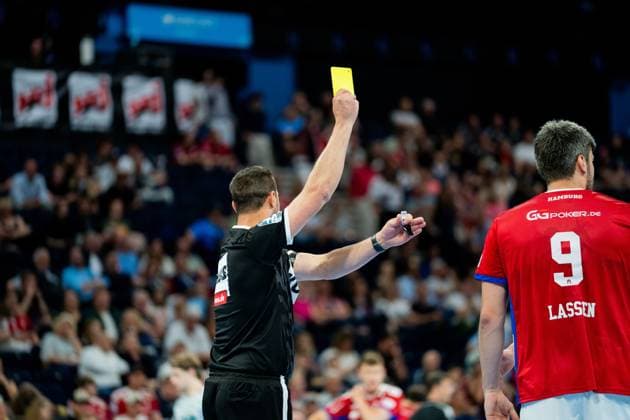 A referee holds up a yellow card during a Bundesliga match between HSV Hamburg and Frisch Auf Göppingen In the background, Jacob Lassen 9 HSV Hamburg stands on the touchline in his red jersey Handball Sport Verein Hamburg vs Frisch Auf Göppingen Copyright: xPhilippHeistx