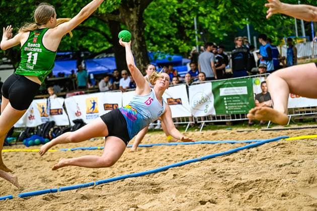 A player 15 throws herself across the sand in flight and attempts a shot on goal while an opponent of Beach Unicorns 11 tries to defend against her In the background, numerous spectators watch the game Spectacular shot attempt 15 in the beach handball game against Beach Unicorns Copyright: xspeedshotx xDorianfrickx