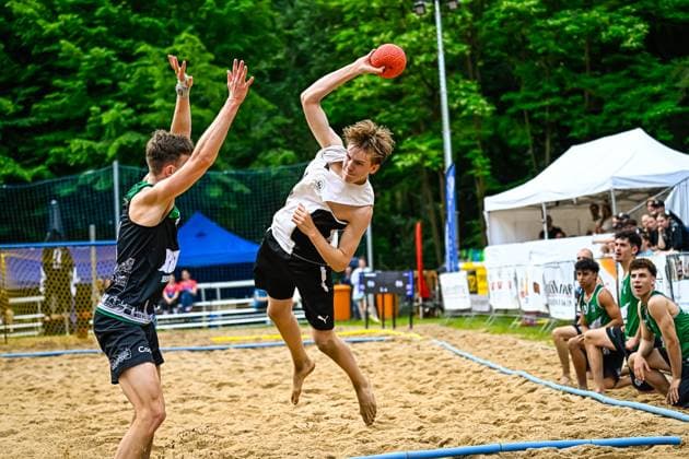 A player in a white jersey jumps up and throws the ball against a defender in a black and green jersey Other players and spectators can be seen on the sidelines in the background Throwing action in beach handball Player jumps against defensive block Copyright: xspeedshotx xDorianfrickx