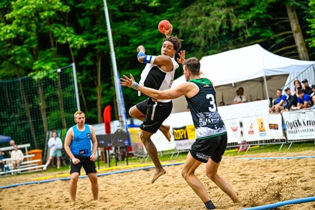 A player in a white jersey jumps up and throws the ball, while player 3 in a black and green jersey with Rotterdam Topsport printed on it tries to block the throw In the background, another player in a blue jersey watches the scene on the sand court Spectacular jump shot at the beach handball tournament Copyright: xspeedshotx xDorianfrickx