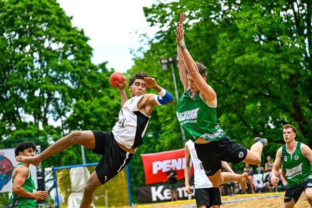 A player in a white jersey jumps to throw and is blocked by an opponent in green The game takes place on an outdoor sand court, sponsor banners of Kempa and Penny are visible in the background Throwing action in beach handball Players in a duel against opponents Copyright: xspeedshotx xDorianfrickx