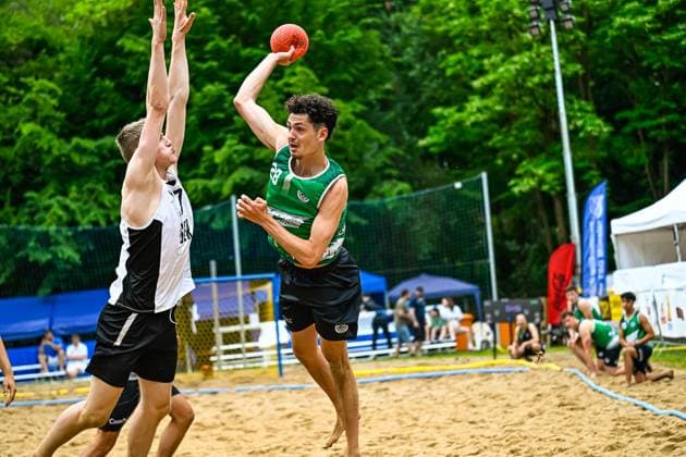 A player in a green jersey 28 jumps up and throws the ball over a defender in a white and black jersey The game takes place on an outdoor sand court, spectators and tents can be seen in the background Throw in beach handball Green attacker against white and black defender Copyright: xspeedshotx xDorianfrickx