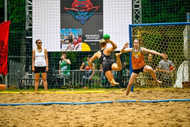 A player with the number 4 jumps in the air and throws the ball at the goal, while goalkeeper 16 spreads her arms out to block the throw In the background, a referee observes the scene on the sand court Player 4 throwing the ball at goal against goalkeeper 16 in beach handball Copyright: xspeedshotx xDorianfrickx