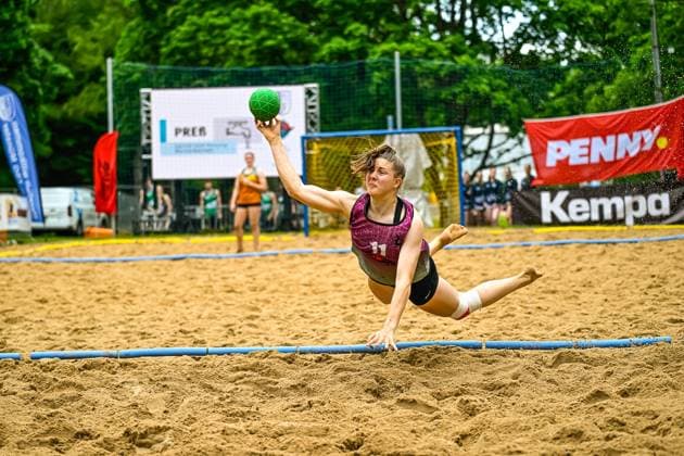 A player with jersey number 11 throws the ball at the goal while falling, stretching horizontally across the sand In the background you can see other players and Penny and Kempa advertising boards Player 11 throwing the ball at the beach handball tournament Copyright: xspeedshotx xDorianfrickx