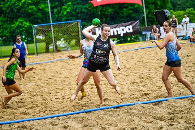 A player with the number 26 in the black jersey of the DMV Group takes a throw and holds the green ball above her head Two opponents in light blue jerseys try to block the throw while another player in the green jersey runs in the background Player 26 DMV Group attempting to throw in beach handball Copyright: xspeedshotx xDorianfrickx