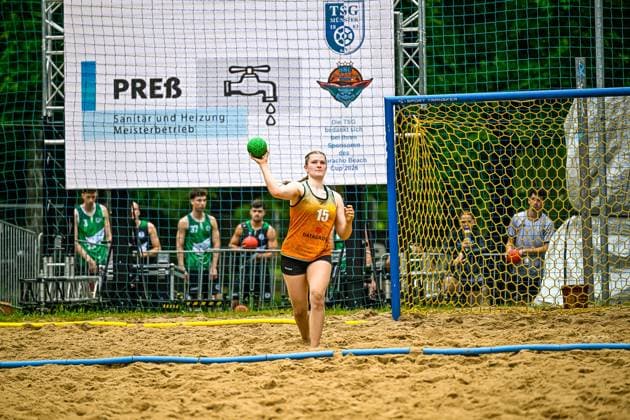 A player with the jersey number 15 and the inscription DATAGROUP takes a throw and holds the green handball in her raised hand In the background you can see players in green jerseys and an advertising banner of the Aracho Beach Cup of TSG Münster Player 15 DATAGROUP throwing in beach handball Copyright: xspeedshotx xDorianfrickx