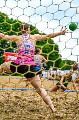 View through the goal net of the German goalkeeper 12 in a pink and white jersey trying to save a green ball with her arm outstretched In the background, other players can be seen in the sand Goalkeeper 12 GER makes a save at the beach handball tournament Copyright: xspeedshotx xDorianfrickx