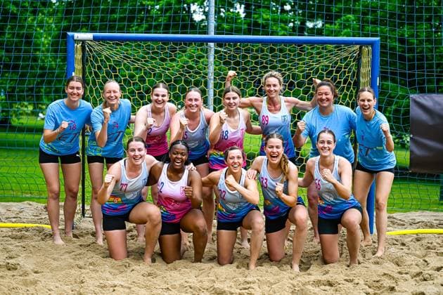A group of players from two teams pose together in front of a blue beach handball goal on sandy ground The players show cheering poses with clenched fists and laugh into the camera Womens team poses cheering in front of the beach handball goal Copyright: xspeedshotx xDorianfrickx