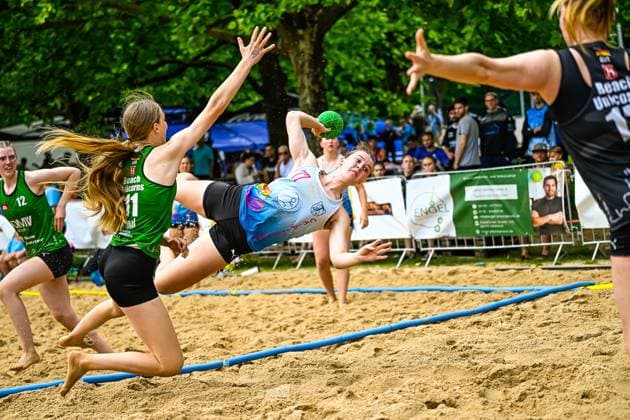 A player in a light blue jersey makes an acrobatic turn and throws herself across the playing field with the ball in her hand Two players from Beach Unicorns 11 and 12 try to block the throw, while 15 of the Beach Unicorns stand ready as goalkeepers Spectacular throw attempt in a beach handball match against Beach Unicorns Copyright: xspeedshotx xDorianfrickx