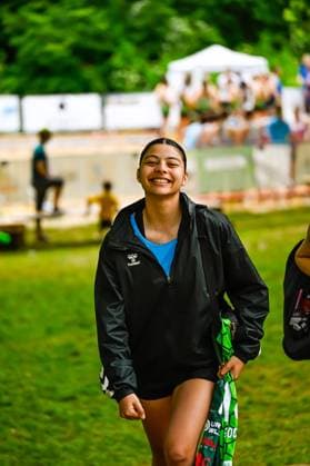 A young player in a black Hummel training jacket and blue undershirt smiling as she runs across a grassy area She is holding a green goalkeeper jersey Smiling player with Hummel jacket at an outdoor sports event Copyright: xspeedshotx xDorianfrickx