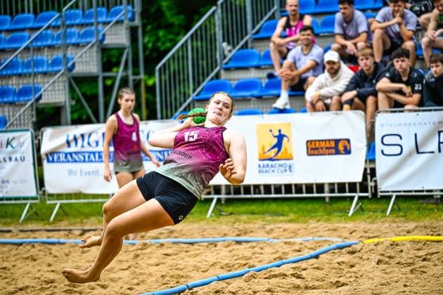 A player with the number 15 in a pink jersey throws at the goal while floating in the air at the Karacho Beach Cup German Beach Open In the background, spectators on blue seats in the stands watch player 15 attempting to throw at the Karacho Beach Cup Copyright: xspeedshotx xDorianfrickx