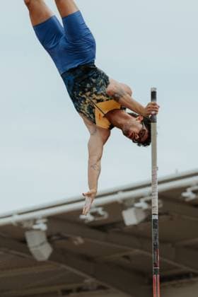 A pole vaulter is in the inverted phase of the jump, holding the pole with both hands while his body hovers upside down over the bar The athlete is wearing a blue top and patterned shorts and can be seen with tattoos on his arms and legs Pole vaulter crossing the bar in competition Copyright: xspeedshotx xBeatricexBauerx